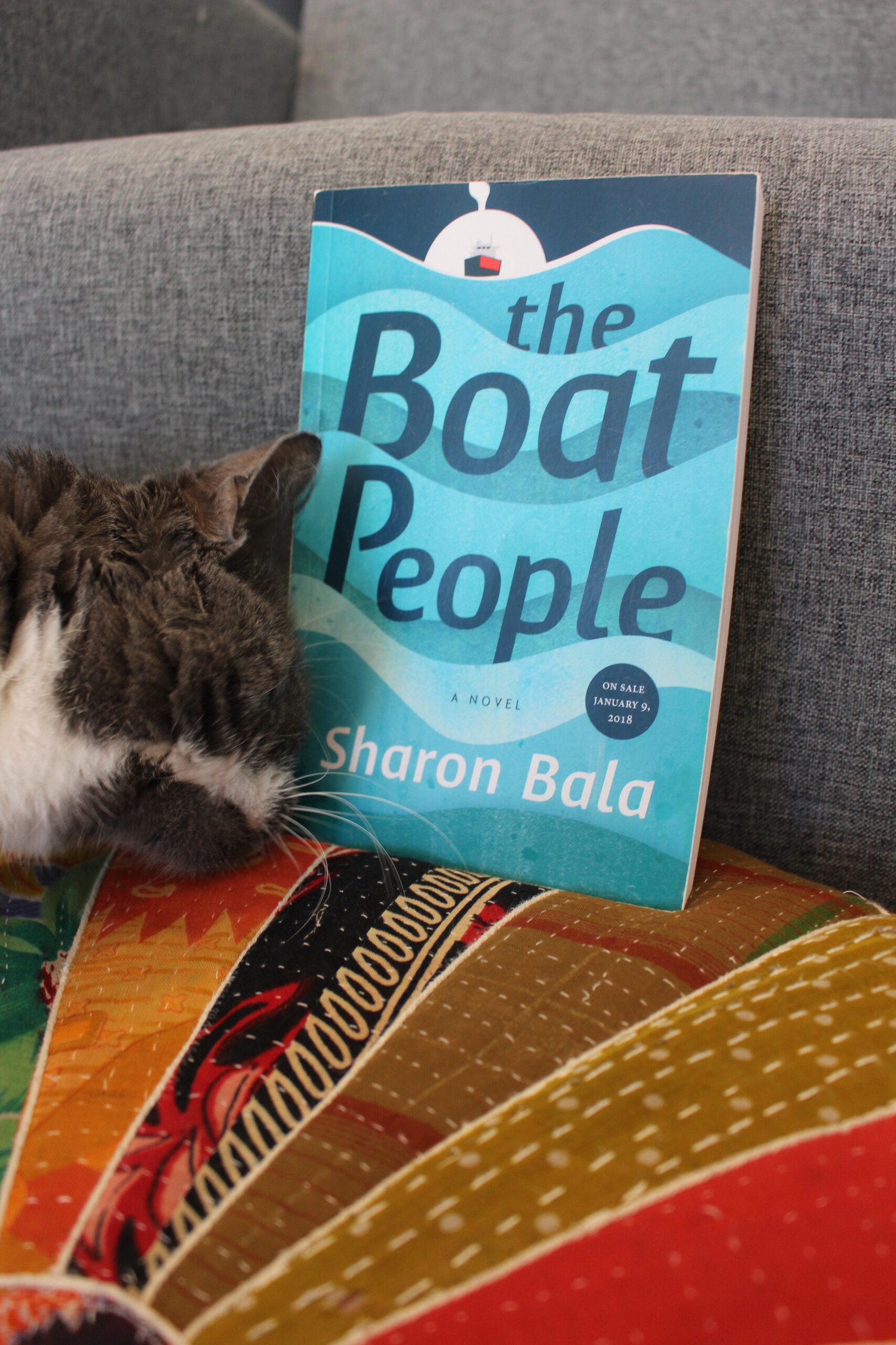 The Boat People by Sharon Bala book pictured on a cushion with a cat's head nuzzling it