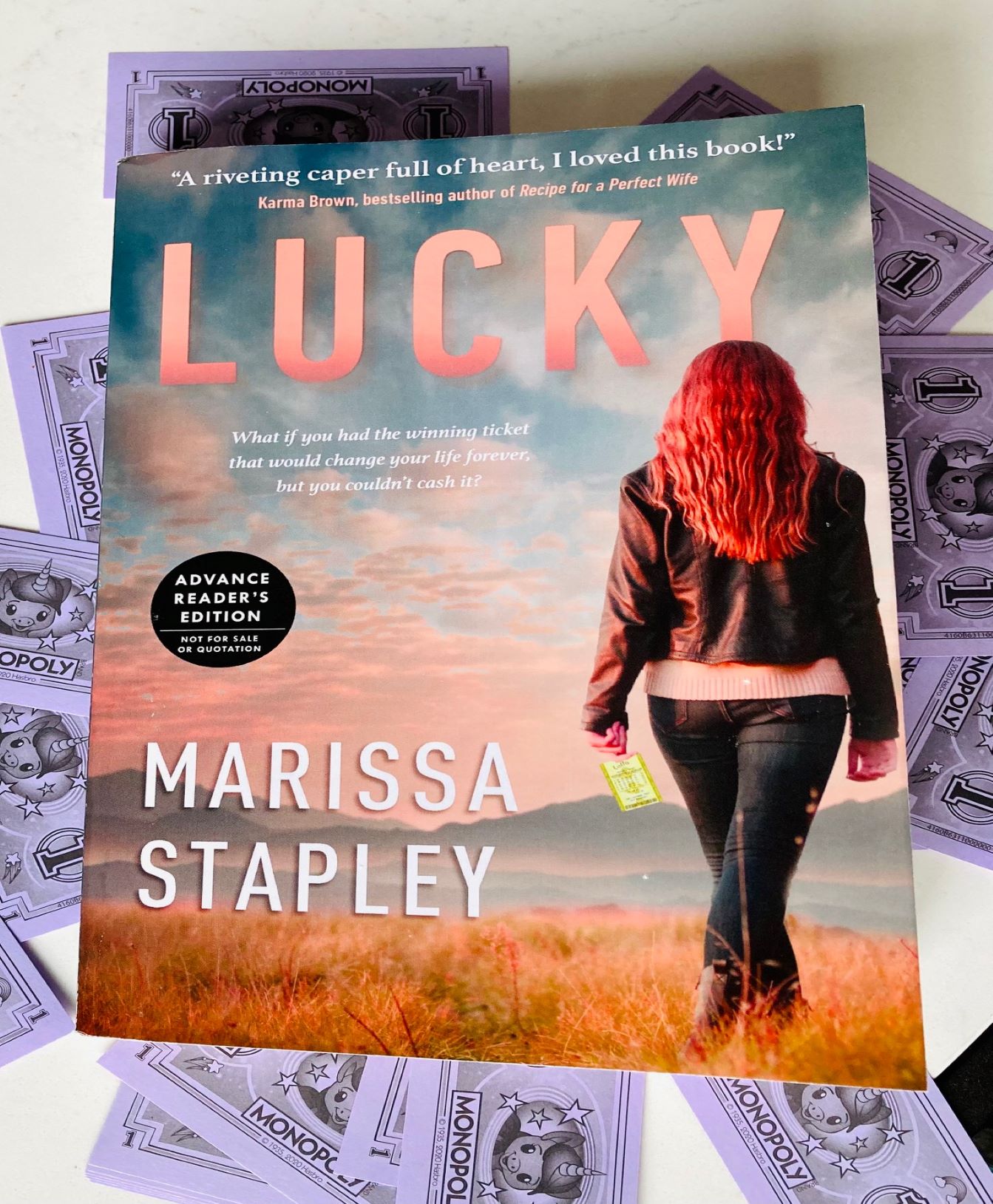 Lucky by Marissa Stapley book on top of purple monopoly play money