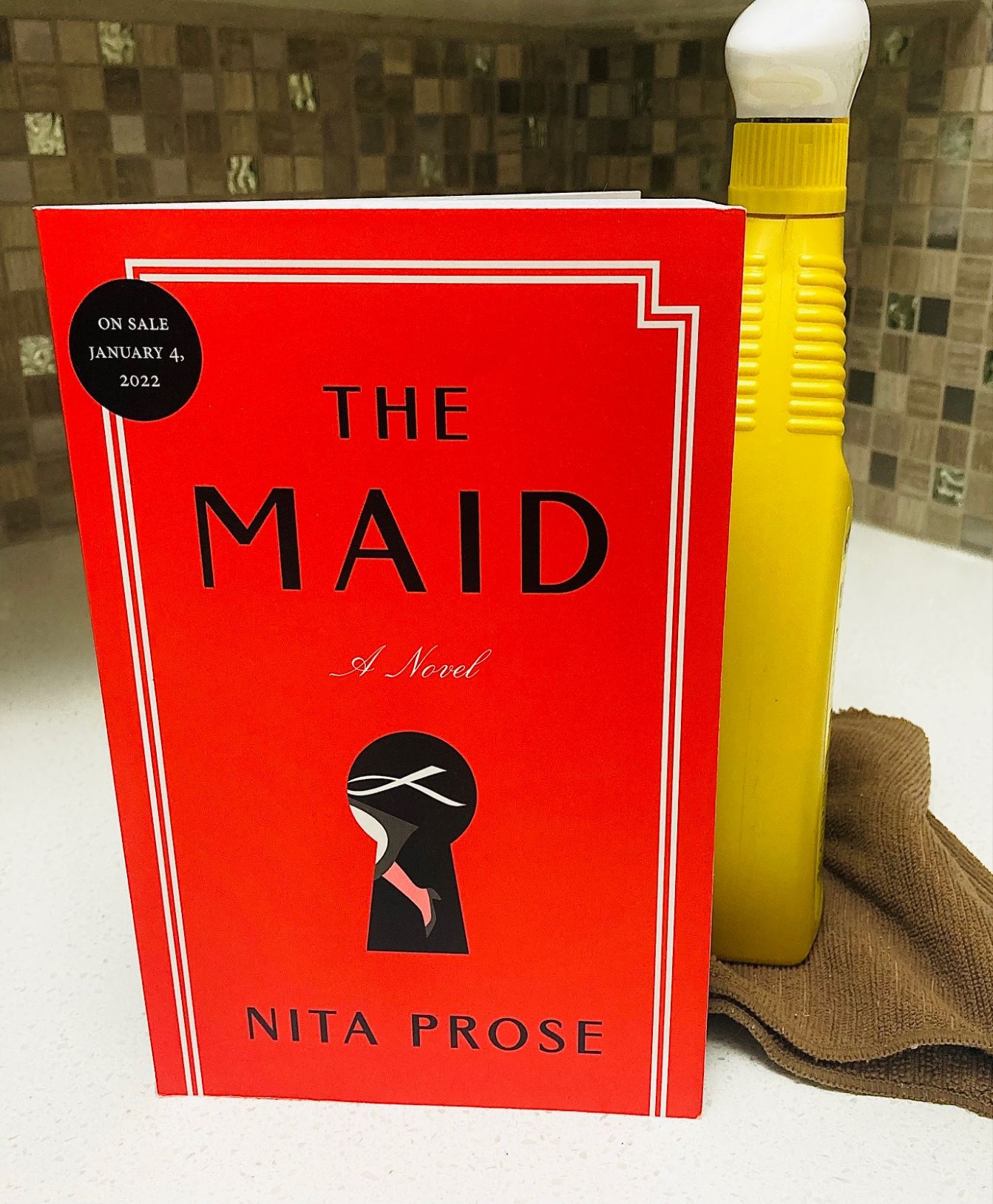 The Maid by Nita Prose book next to a yellow spray bottle and brown cleaning cloth on a white countertop