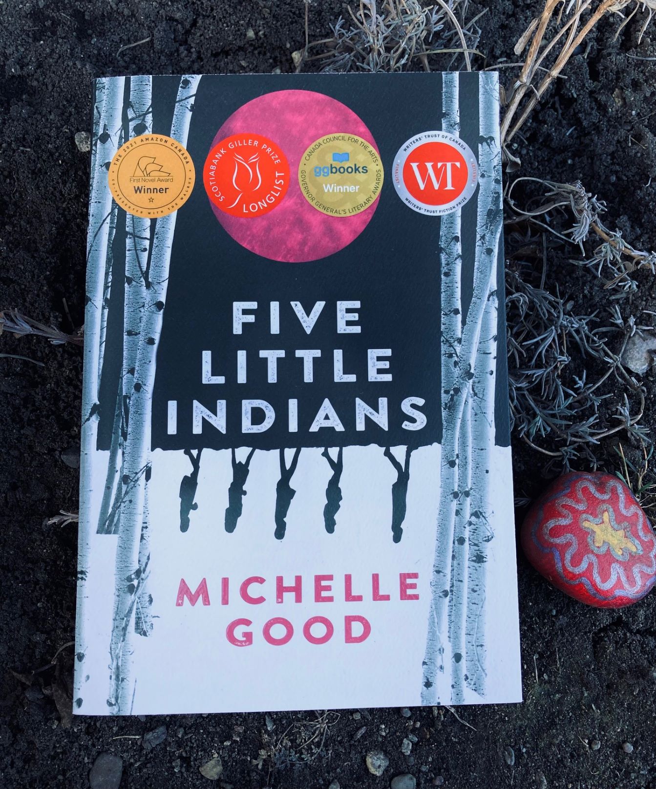 picture of Five Little Indians book by Michelle Good on the ground with a painted rock and dead branches beneath it