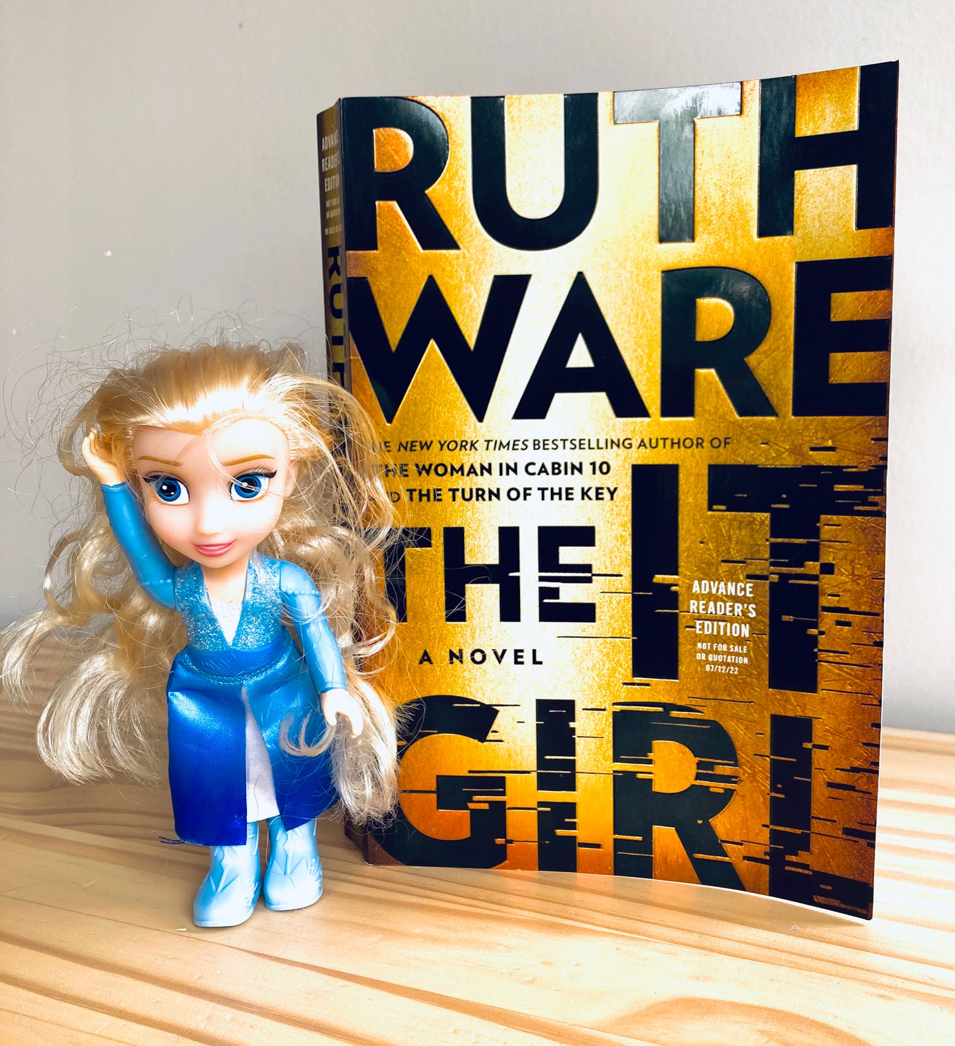 picture of The It Girl book by Ruth Ware, next to a small doll of a blond haired girl in a blue dress