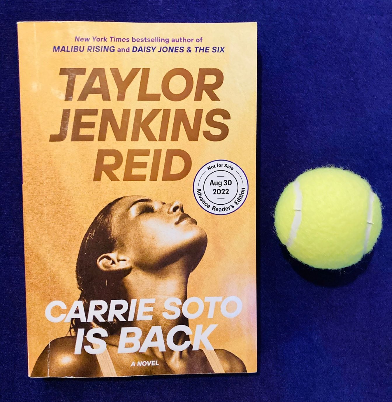 picture of Carrie Soto is Back by Taylor Jenkins Reid, on a purple background with a yellow tennis ball beside the book