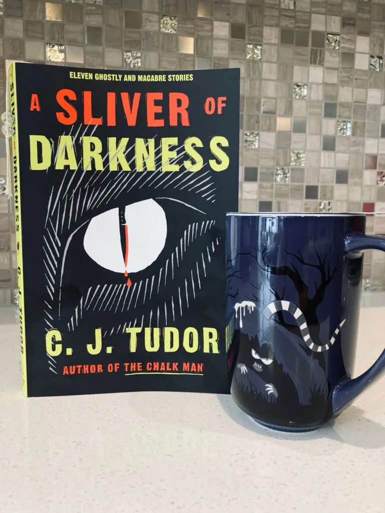 A Sliver of Darkness by C.J. Tudor book pictured beside a mug with creepy eyes and a scary looking snake on it