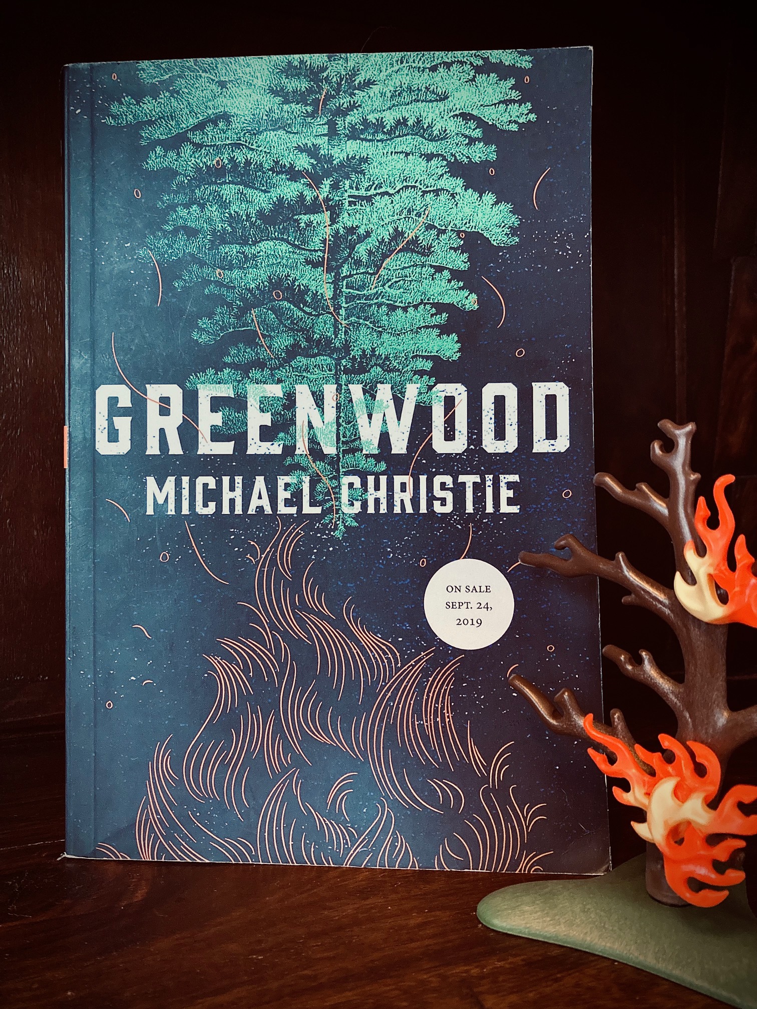 picture of Greenwood by Michael Christie book, next to a small plastic tree with flames on it