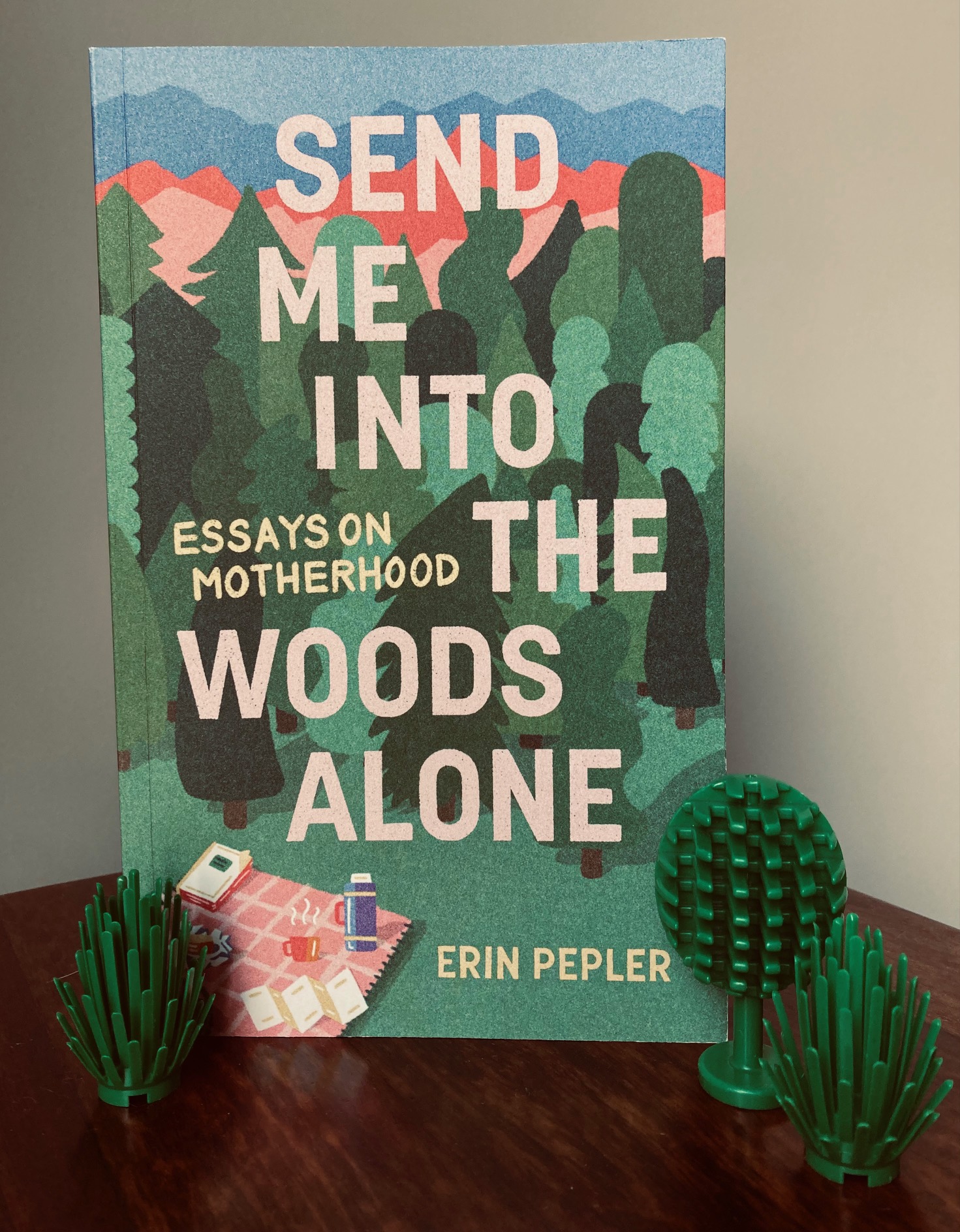 picture of book titled Send Me Into the Woods Alone by Erin Pepler, with some small green plastic trees in front of the cover along the bottom of the book, on a gray background