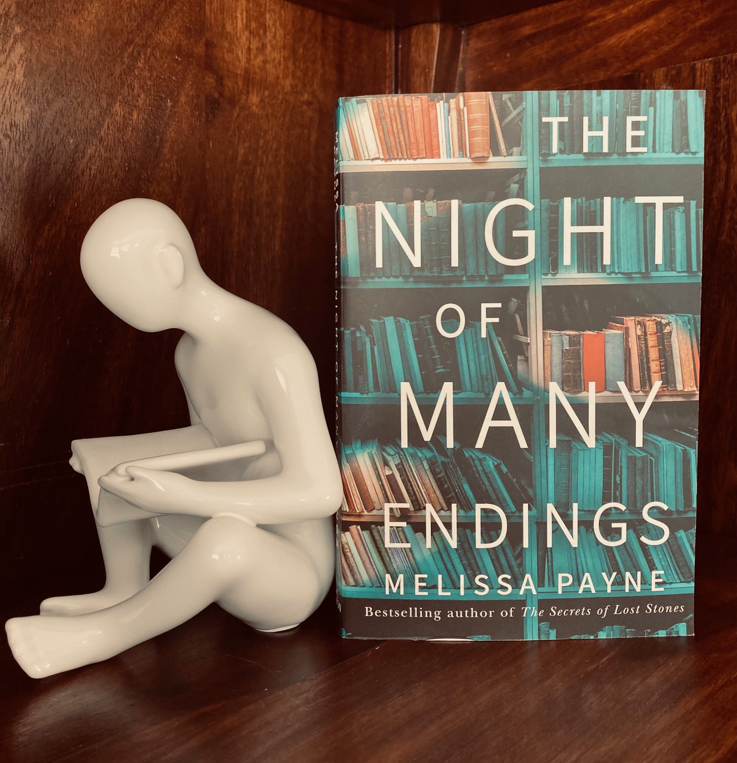 The Night of Many Endings by Melissa Payne book next to a book end of a white figure sitting down and reading a book