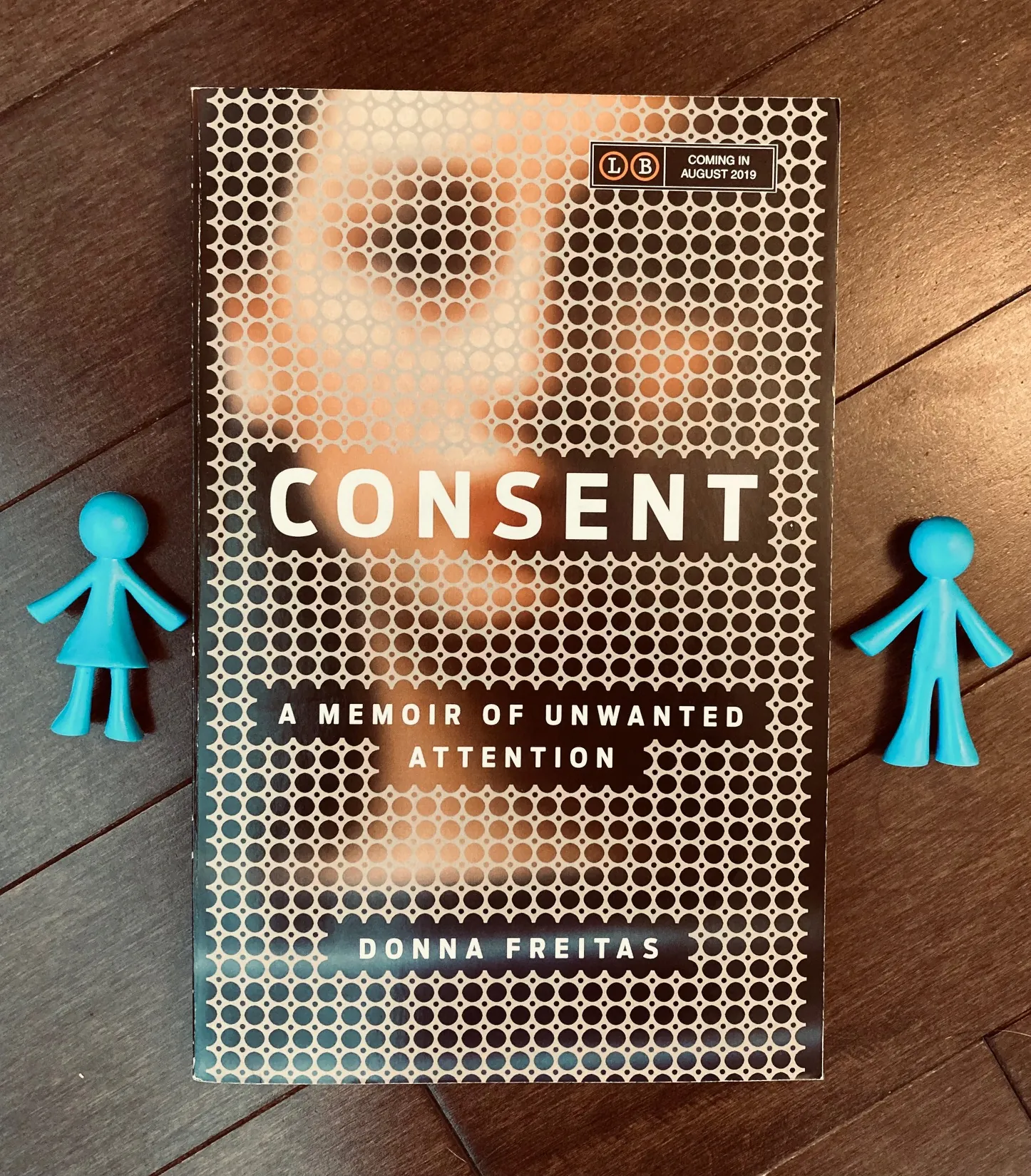 Consent by Donna Freitas book with a blue plastic girl and boy figure on either side of the book, lying on top of a wooden background
