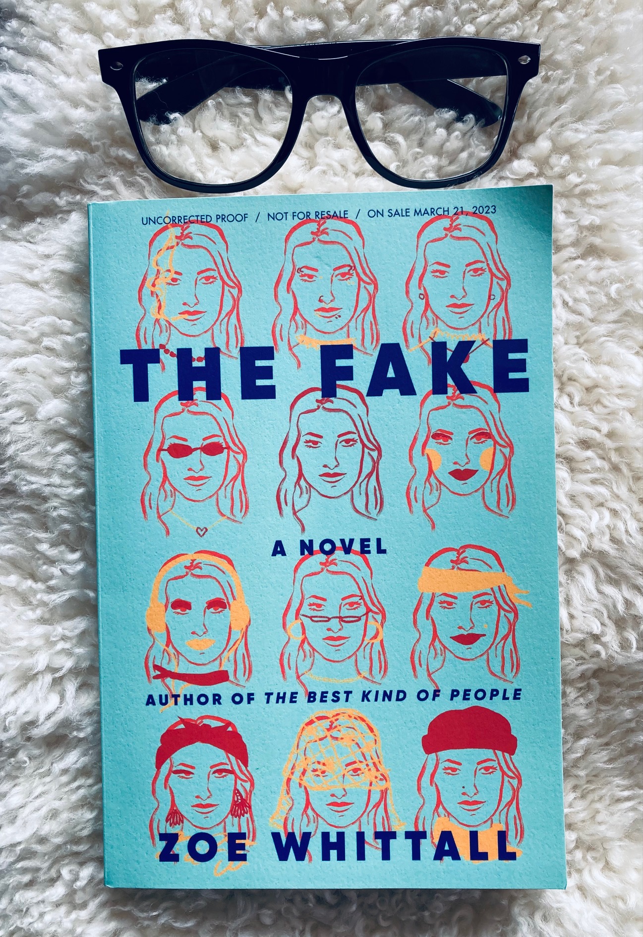 The Fake by Zoe Whittall book pictured on a white shag background with a pair of black plastic glasses frames above the book