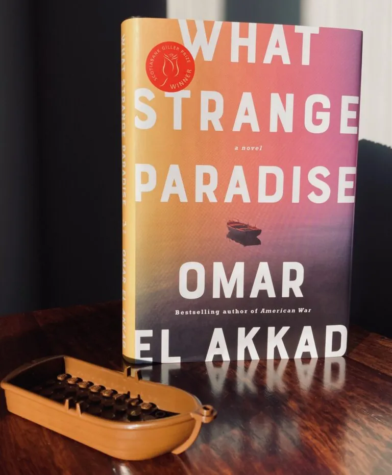 What Strange Paradise by Omar El Akkad book pictured on a wooden surface with a small brown lego boat in front of it