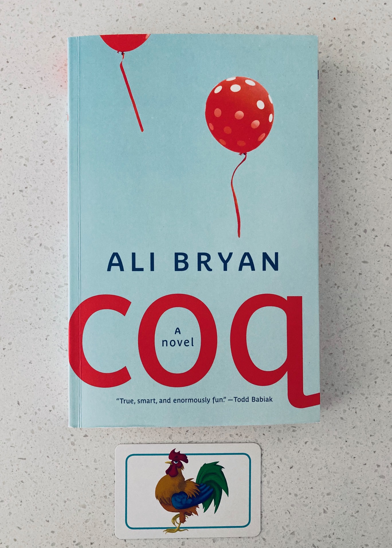 Coq by Ali Bryan book pictured on a white background with a small picture of a rooster underneath it