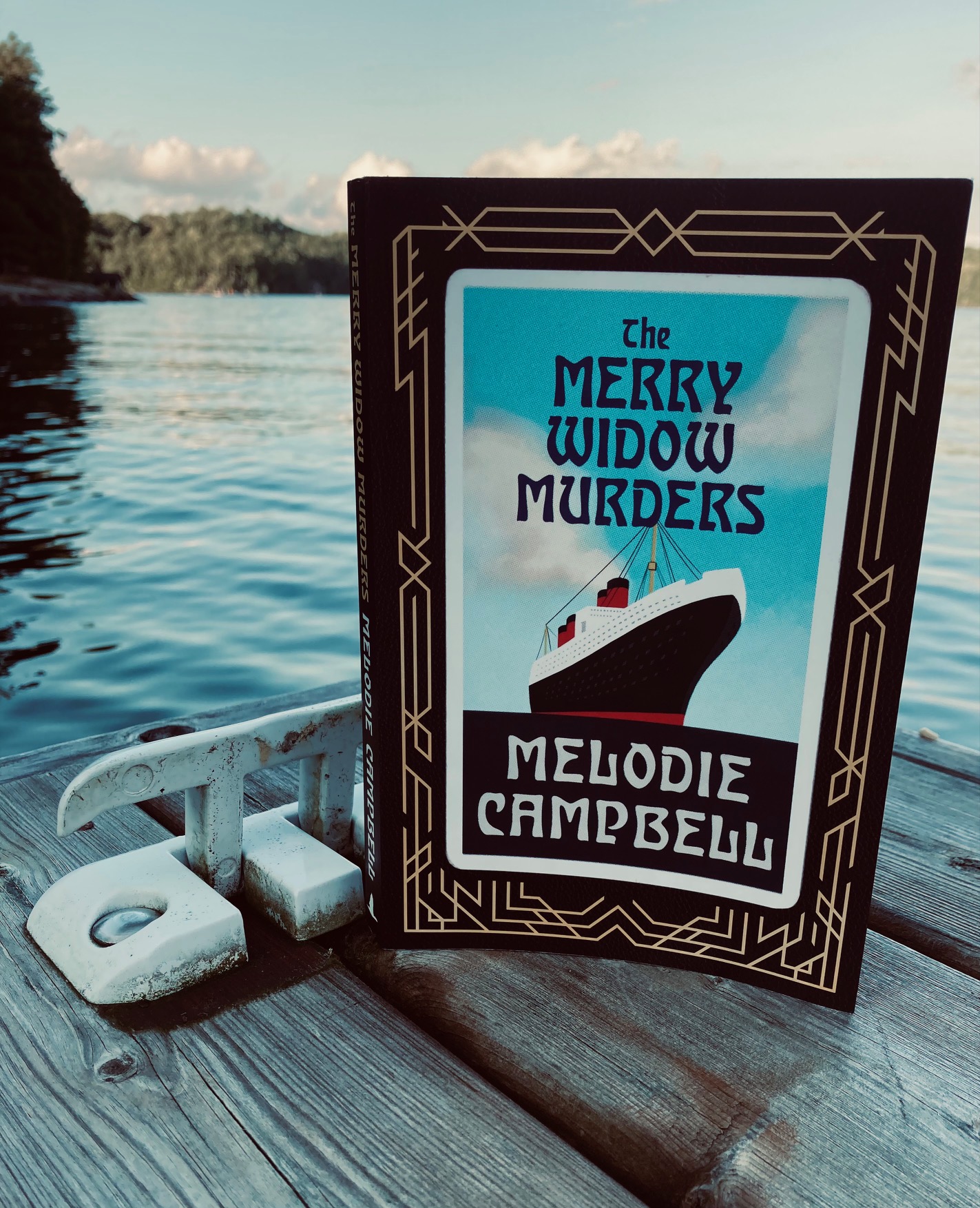 The Merry Widow Murders by Melodie Campbell book pictured on a dock in front of a lake