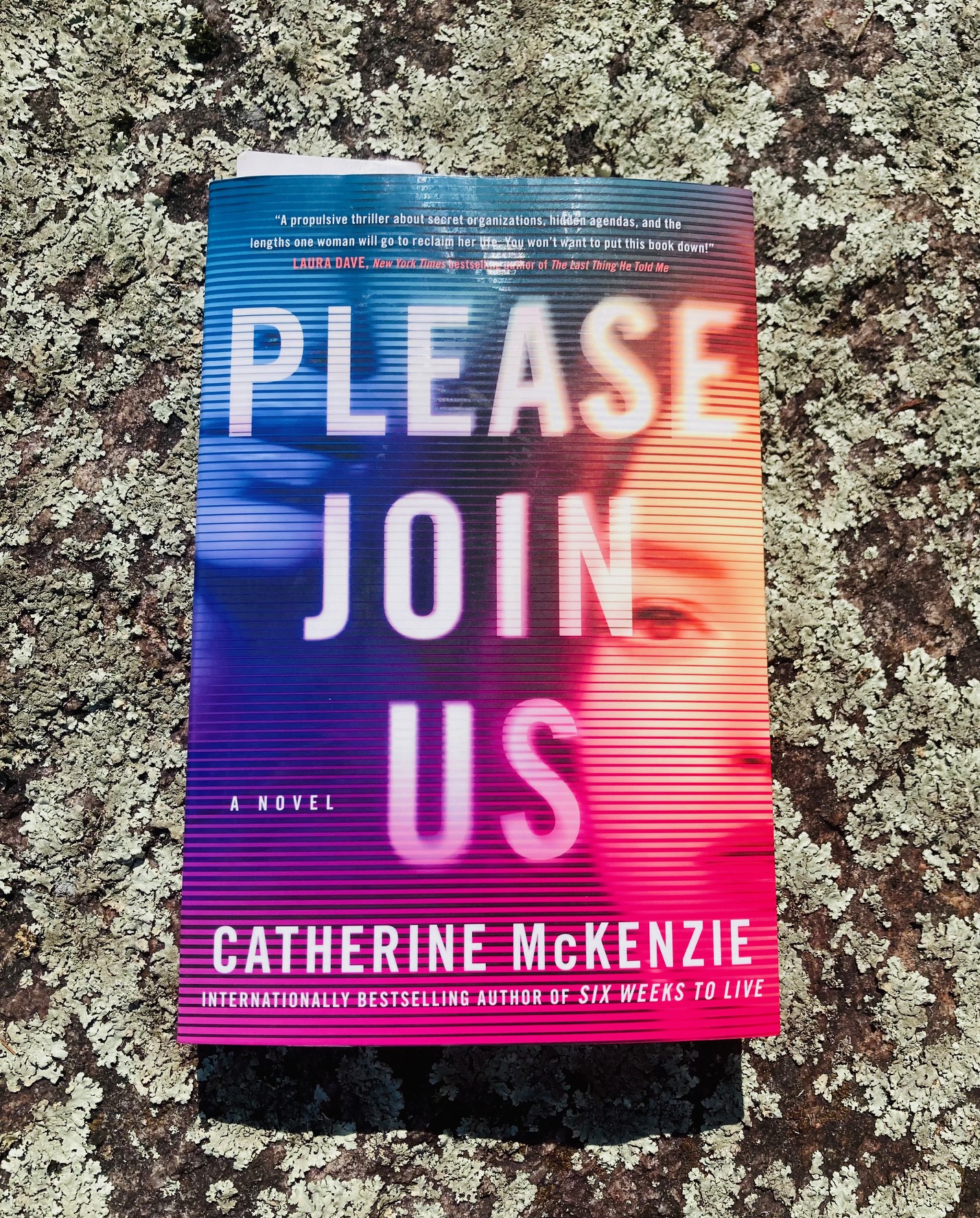 Please Join Us by Catherine McKenzie book pictured on a lichen covered rock